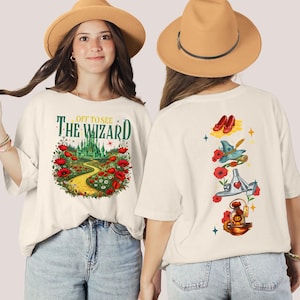 May include: Cream-colored t-shirt with a Wizard of Oz theme. The front features the text "Off to See The Wizard" above a colorful illustration of the yellow brick road, poppies, and the Emerald City. The back has illustrations of ruby slippers, a witch's hat, and other iconic imagery.