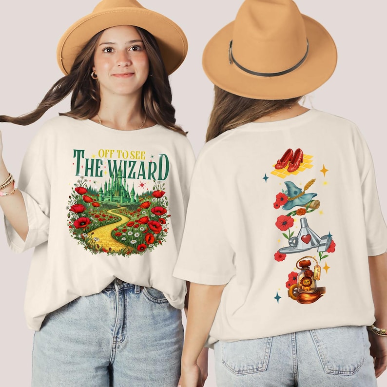 May include: Cream-colored t-shirt with a Wizard of Oz theme. The front features the text "Off to See The Wizard" above a colorful illustration of the yellow brick road, poppies, and the Emerald City. The back has illustrations of ruby slippers, a witch's hat, and other iconic imagery.