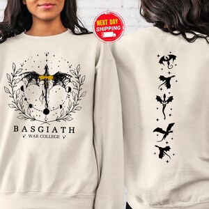 May include: Cream-colored sweatshirt with a black and gold dragon design on the front and back. The front features a dragon with the text "BASGIATH WAR COLLEGE". The back has five black dragon silhouettes.