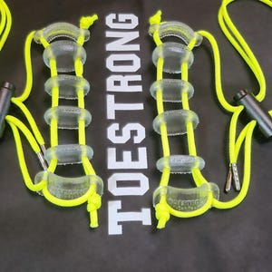 May include: Neon yellow skipping rope with clear plastic handles and black cylindrical handles. The text "TOESTRONG" is printed in white on a black background. The skipping rope is designed for fitness and exercise.