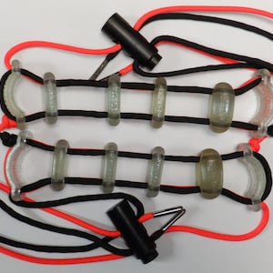 Pode incluir: Dois dispositivos de tração paralelos com cordões pretos e laranja néon enfiados através de segmentos de plástico translúcido, texturizado e semicircular. Cada unidade inclui um componente de tensão cilíndrico preto e um gancho de metal.