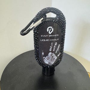 May include: Black liquid chalk bottle with a handprint design. The bottle is embellished with black rhinestones and a silver carabiner clip. The text "EVMT BRANDS LIQUID CHALK" is printed on the bottle.