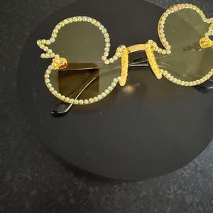 May include: Gold-rimmed sunglasses in a Mickey Mouse shape, adorned with small, iridescent rhinestones. The lenses are light yellow, and the bridge and arms are gold. A fun, novelty eyewear item.