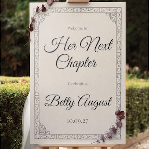 May include: A beige sign on an easel with the text "Welcome to Her Next Chapter Celebrating Betty August 10.09.27" in elegant script. The sign has a decorative border and is adorned with floral accents.