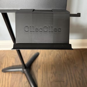 May include: A black music stand with the text "OllecOllec" embossed on the front. The stand has a three-legged base and a tray to hold sheet music. The stand is made of metal and is designed to be adjustable.