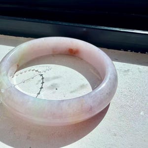 May include: A smooth, polished bangle bracelet crafted from translucent light pink and white stone. The circular band features subtle, natural inclusions of pale green and small, distinct reddish-orange spots distributed across its surface.