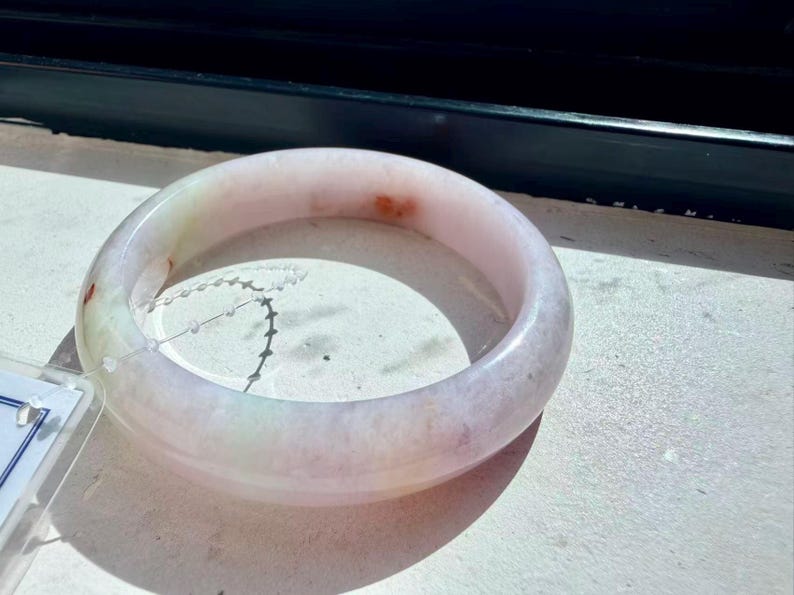 May include: A smooth, polished bangle bracelet crafted from translucent light pink and white stone. The circular band features subtle, natural inclusions of pale green and small, distinct reddish-orange spots distributed across its surface.