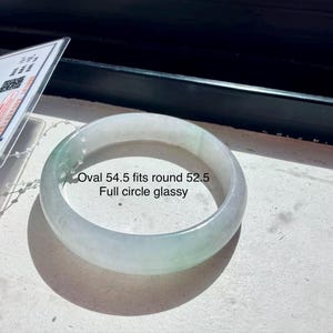 May include: A glassy, pale green jade bangle bracelet with a smooth, rounded surface. The text on the image reads: Oval 54.5 fits round 52.5 Full circle glassy. A white identification tag with a QR code is attached to the bangle by a string.
