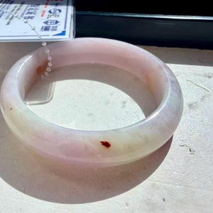 May include: A smooth, translucent pale pink and white jade bangle bracelet with a polished finish. The circular jewelry piece features subtle natural color variations and a single small, distinct reddish-brown inclusion on its outer surface.