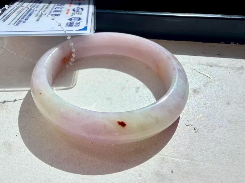 May include: A smooth, translucent pale pink and white jade bangle bracelet with a polished finish. The circular jewelry piece features subtle natural color variations and a single small, distinct reddish-brown inclusion on its outer surface.