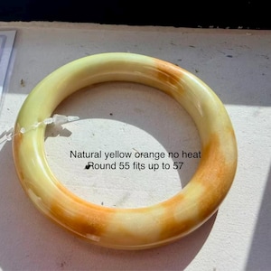 May include: A smooth, round bangle bracelet featuring a natural yellow and orange colour palette. The text on the surface reads: Natural yellow orange no heat, Round 55 fits up to 57. The bangle has a polished finish and a solid, circular form.