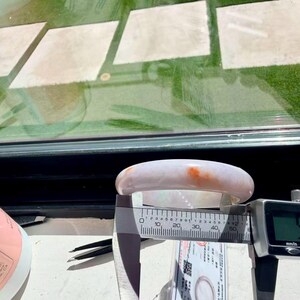 May include: A smooth, pale lavender jade bangle bracelet with a distinct orange-red inclusion. A digital caliper is positioned across the inner diameter of the bangle, displaying a measurement of 57.0 millimeters on its clear digital screen.