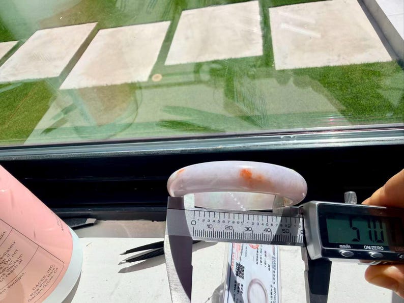 May include: A smooth, pale lavender jade bangle bracelet with a distinct orange-red inclusion. A digital caliper is positioned across the inner diameter of the bangle, displaying a measurement of 57.0 millimeters on its clear digital screen.