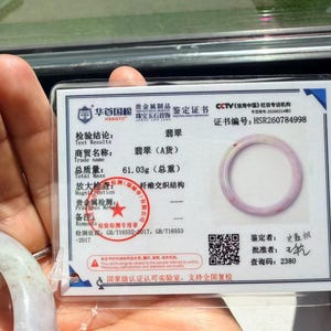 May include: A jade bangle certificate of authenticity featuring a photograph of a pale lavender and white jade bangle. The document displays the certificate number HSR260784998, a total mass of 61.03g, a red circular stamp, and a QR code for verification.