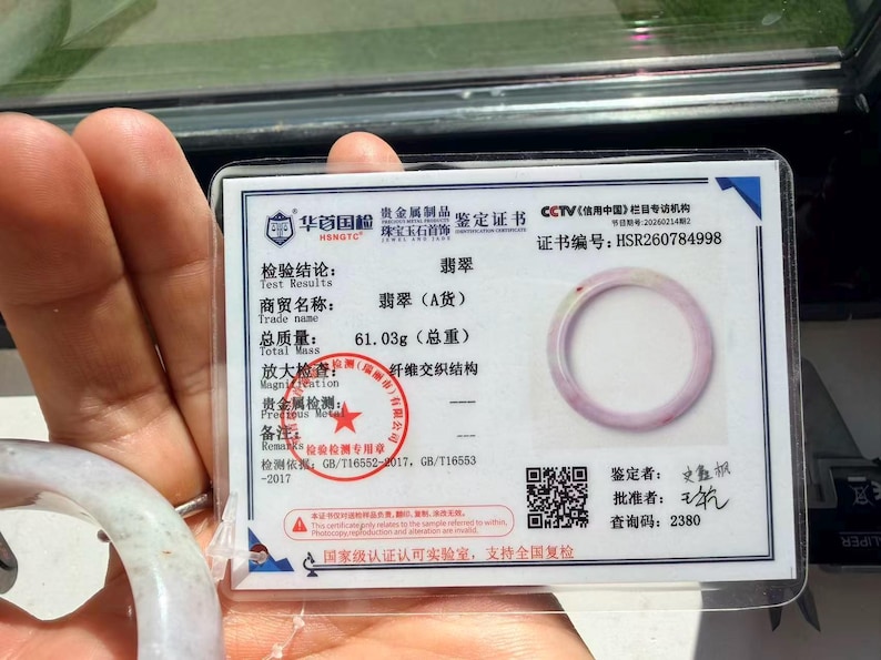May include: A jade bangle certificate of authenticity featuring a photograph of a pale lavender and white jade bangle. The document displays the certificate number HSR260784998, a total mass of 61.03g, a red circular stamp, and a QR code for verification.