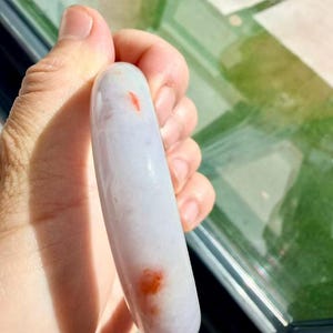 May include: A smooth, rounded bangle bracelet crafted from translucent, pale lavender stone. The surface of the bangle features distinct, small, vibrant orange-red inclusions scattered along its polished, curved exterior profile.