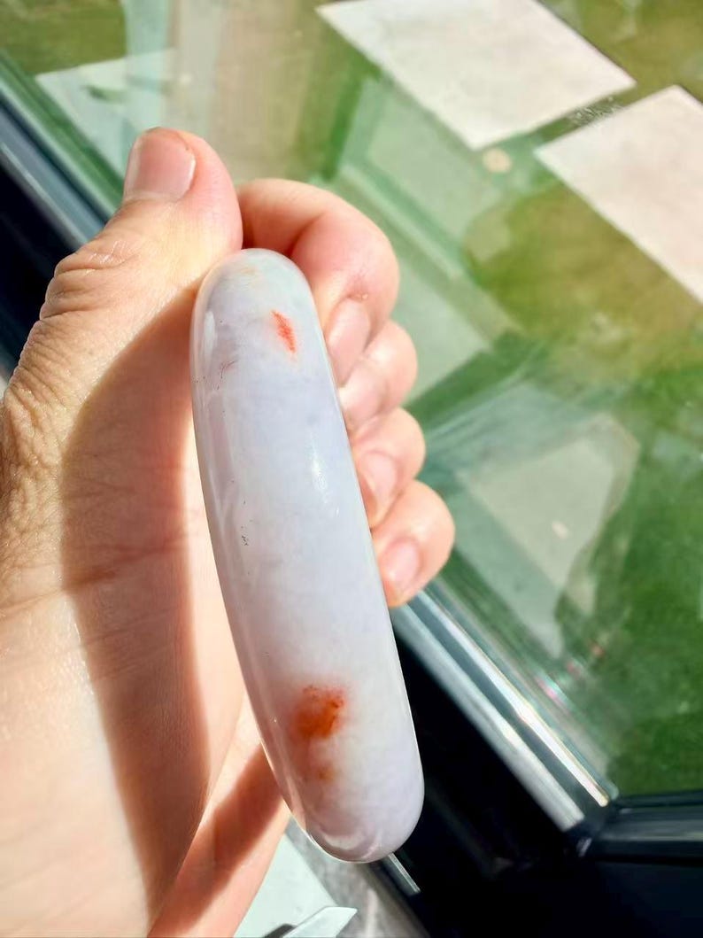 May include: A smooth, rounded bangle bracelet crafted from translucent, pale lavender stone. The surface of the bangle features distinct, small, vibrant orange-red inclusions scattered along its polished, curved exterior profile.