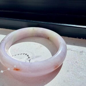 May include: A smooth, circular bangle bracelet crafted from translucent stone with a pale lavender hue. The surface features subtle, cloudy white inclusions and small, distinct reddish-orange mineral deposits scattered along the rounded band.