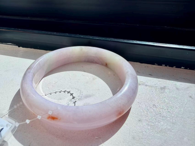 May include: A smooth, circular bangle bracelet crafted from translucent stone with a pale lavender hue. The surface features subtle, cloudy white inclusions and small, distinct reddish-orange mineral deposits scattered along the rounded band.