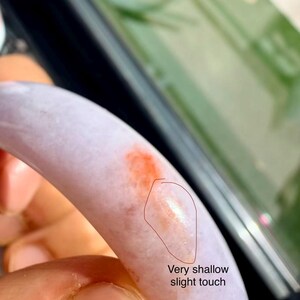 May include: A smooth, lavender-colored bangle bracelet featuring a distinct orange-red patch and a small, circled area labeled with the text Very shallow slight touch, indicating a minor surface imperfection on the polished stone material.