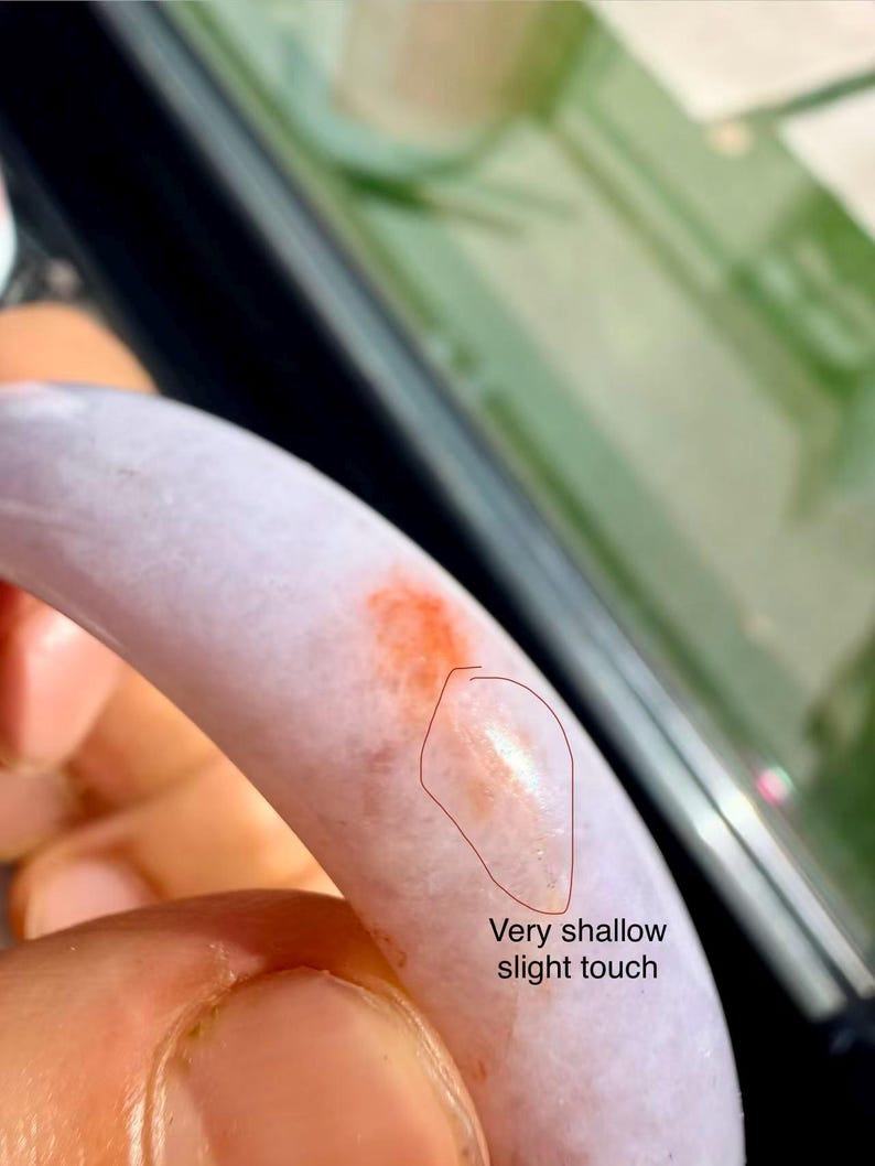 May include: A smooth, lavender-colored bangle bracelet featuring a distinct orange-red patch and a small, circled area labeled with the text Very shallow slight touch, indicating a minor surface imperfection on the polished stone material.