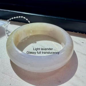 May include: A light lavender bangle bracelet with a smooth, rounded surface and glassy full translucency. The bracelet features a thin, clear plastic string attached to the top edge and includes the text Light lavender Glassy full translucency.