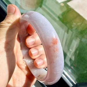 May include: A smooth, polished lavender jade bangle bracelet with a rounded profile. The stone features a translucent, pale purple base color with a distinct, small patch of reddish-orange mineral inclusion visible on the outer surface.