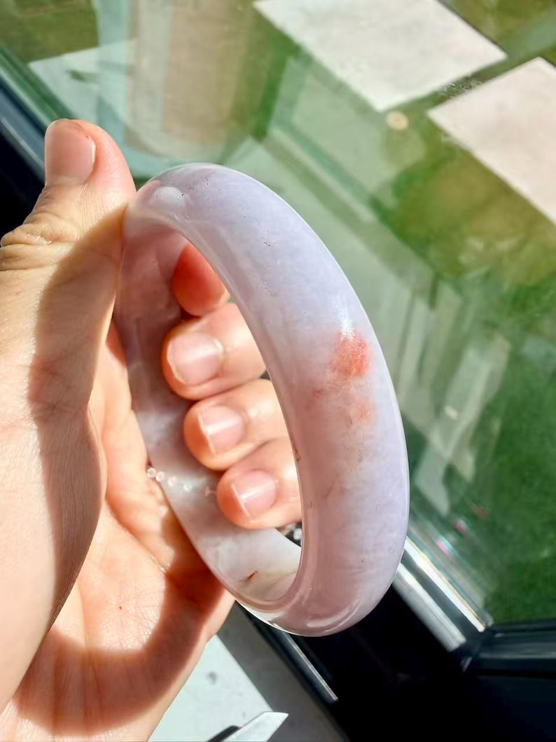 May include: A smooth, polished lavender jade bangle bracelet with a rounded profile. The stone features a translucent, pale purple base color with a distinct, small patch of reddish-orange mineral inclusion visible on the outer surface.