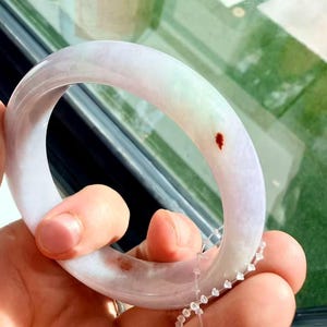 May include: A smooth, polished jade bangle bracelet featuring a translucent lavender and pale green color palette. The circular band displays distinct natural reddish-brown inclusions and is accompanied by a small, clear plastic security tag.