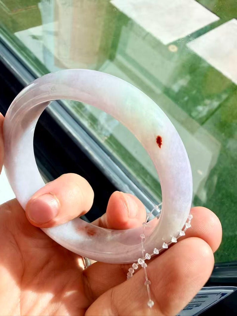 May include: A smooth, polished jade bangle bracelet featuring a translucent lavender and pale green color palette. The circular band displays distinct natural reddish-brown inclusions and is accompanied by a small, clear plastic security tag.