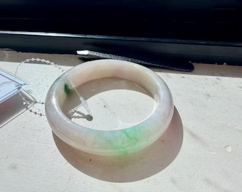 May include: A smooth, circular bangle bracelet crafted from translucent, off-white stone with subtle, mottled green inclusions. The bracelet features a thin, clear plastic security tag attached with a small, beaded cord loop.