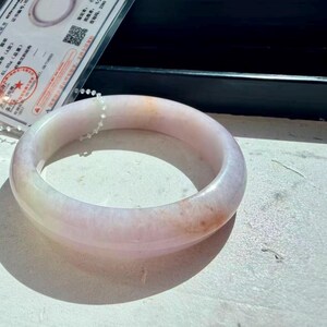 May include: A smooth, polished bangle bracelet crafted from translucent stone with a pale lavender and white base color, featuring subtle hints of soft orange and peach marbling throughout the circular, rounded band structure.