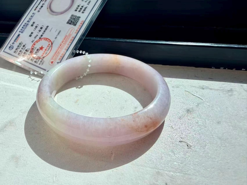 May include: A smooth, polished bangle bracelet crafted from translucent stone with a pale lavender and white base color, featuring subtle hints of soft orange and peach marbling throughout the circular, rounded band structure.