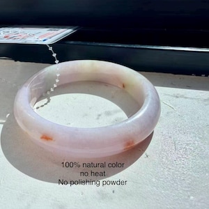 May include: A smooth, translucent, pale lavender and white jade bangle bracelet with subtle orange-red inclusions. The bracelet features a plastic tag attached by a white cord and text printed below it stating 100% natural color, no heat, no polishing powder.