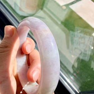 May include: A smooth, polished lavender and white jade bangle bracelet with a rounded profile. The stone features natural cloudy inclusions and subtle color variations throughout the surface, showcasing a translucent quality under bright light.