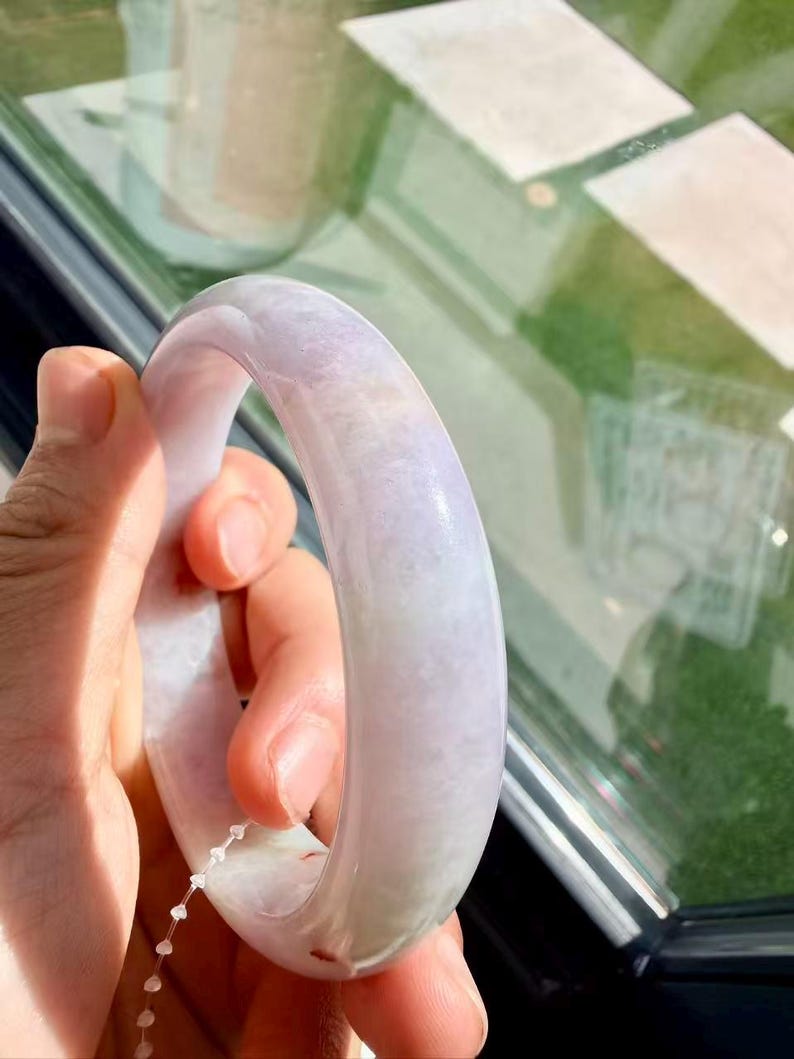 May include: A smooth, polished lavender and white jade bangle bracelet with a rounded profile. The stone features natural cloudy inclusions and subtle color variations throughout the surface, showcasing a translucent quality under bright light.