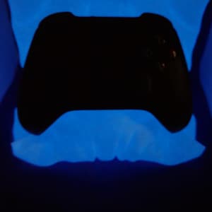 May include: A black video game controller is centered in the image, surrounded by a glowing blue light. The controller's buttons and joysticks are visible. The background is dark, enhancing the contrast of the controller and the blue light.
