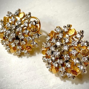 Pode incluir: Um par de brincos redondos com um aglomerado de pedras claras e brilhantes cravadas numa base de metal dourado. O design incorpora pequenos discos dourados texturizados entre as pedras, criando uma aparência floral densa e multicamadas.