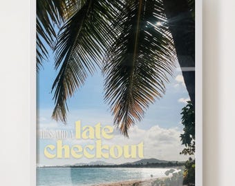 Op de afbeelding: Ingelijste print met een tropische strandscène met palmtakken, turquoise water en een zandstrand. De tekst "THIS AND A late checkout" wordt in een gele lettertype over de afbeelding weergegeven. De lucht is blauw met wat wolken.