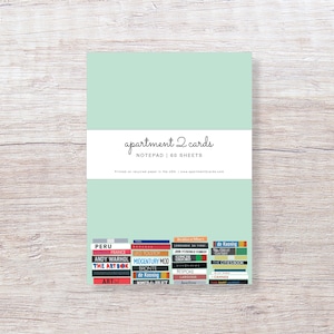 May include: A light green notepad with the text "apartment 2 cards" and "NOTEPAD | 60 SHEETS" printed on the front. The back of the notepad features a collage of book covers with various titles and authors.