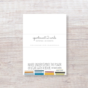 May include: A white notepad with a quote from Ruth Bader Ginsburg: "Never underestimate the power of a girl with a book." The notepad is decorated with a stack of colorful books. The notepad is labeled "apartment 2 cards" and "Notepad | 60 sheets".