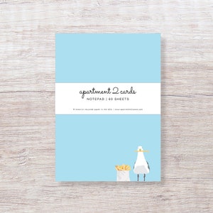 May include: A light blue notepad with a white label that says "apartment 2 cards" and "NOTEPAD | 60 SHEETS". The notepad has a cartoon illustration of a seagull holding a stick in its beak and a bag of french fries.