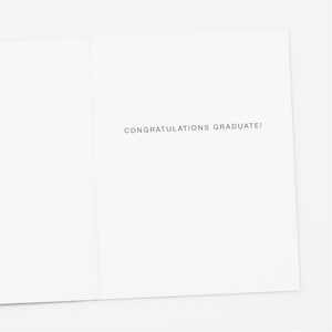 Op de afbeelding: Een witte wenskaart met de tekst "CONGRATULATIONS GRADUATE!" in zwarte letters gedrukt. De kaart is open, waardoor de binnenkant en de tekst zichtbaar zijn. De kaart is blanco, met een eenvoudig en elegant ontwerp.