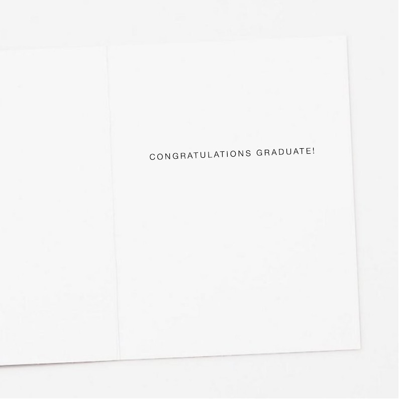Op de afbeelding: Een witte wenskaart met de tekst "CONGRATULATIONS GRADUATE!" in zwarte letters gedrukt. De kaart is open, waardoor de binnenkant en de tekst zichtbaar zijn. De kaart is blanco, met een eenvoudig en elegant ontwerp.