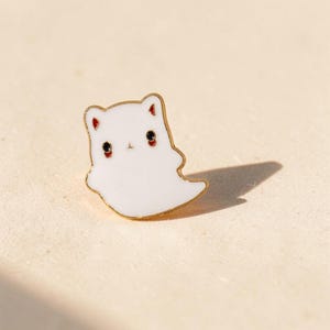 May include: A white enamel pin shaped like a small ghost with gold-toned metal edges. The ghost features two black circular eyes with red accents, small red-lined ears, and a simple, rounded body silhouette with a pointed tail end.