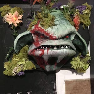 May include: A grotesque goblin mask with green skin and red blood-like markings. The mask is adorned with moss, flowers, and foliage. The goblin has sharp teeth and is mounted on a black surface.
