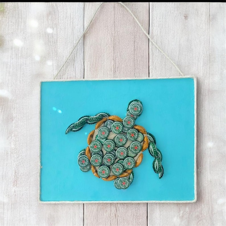 以下が含まれることがあります： 海亀のデザインが施された額入りの壁掛けアート。カメは緑と金の王冠でできており、水色の背景に配置されています。フレームは白で、アートワークは紐から吊り下げられています。