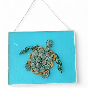 以下が含まれることがあります： ハイネケンのボトルキャップで作られたカメのデザインの手作りの壁の芸術作品。カメは水色の背景に配置され、白いロープで縁取られています。このアートワークはすぐに飾ることができます。