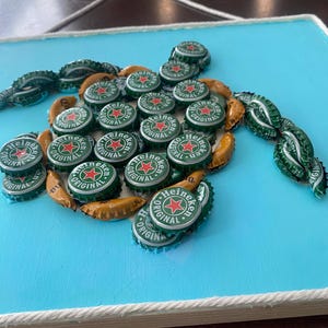 以下が含まれることがあります： ハイネケンのボトルキャップで作られたカメのモザイク。薄い青色の背景に配置されています。カメの体は緑と金のボトルキャップで形作られ、ハイネケンのロゴが見えます。作品は白いロープで縁取られています。