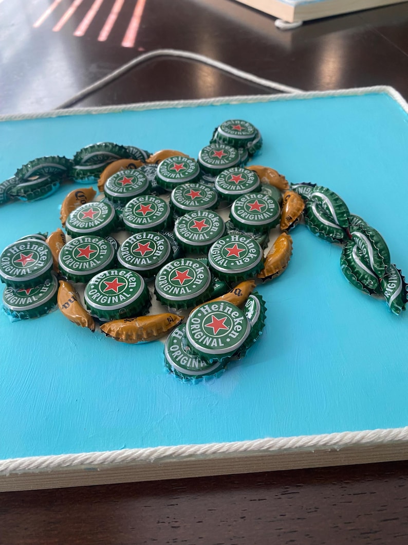 以下が含まれることがあります： ハイネケンのボトルキャップで作られたカメのモザイク。薄い青色の背景に配置されています。カメの体は緑と金のボトルキャップで形作られ、ハイネケンのロゴが見えます。作品は白いロープで縁取られています。